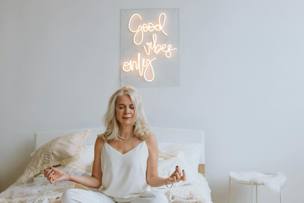 White woman meditating in minimalist room with a "Good Vibes Only" sign — representing stereotypical wellness culture and surface-level positivity messaging.
