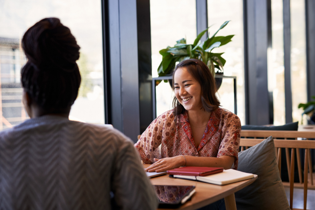 Two women sitting together with coffee and notebooks, sharing a relaxed, natural conversation.